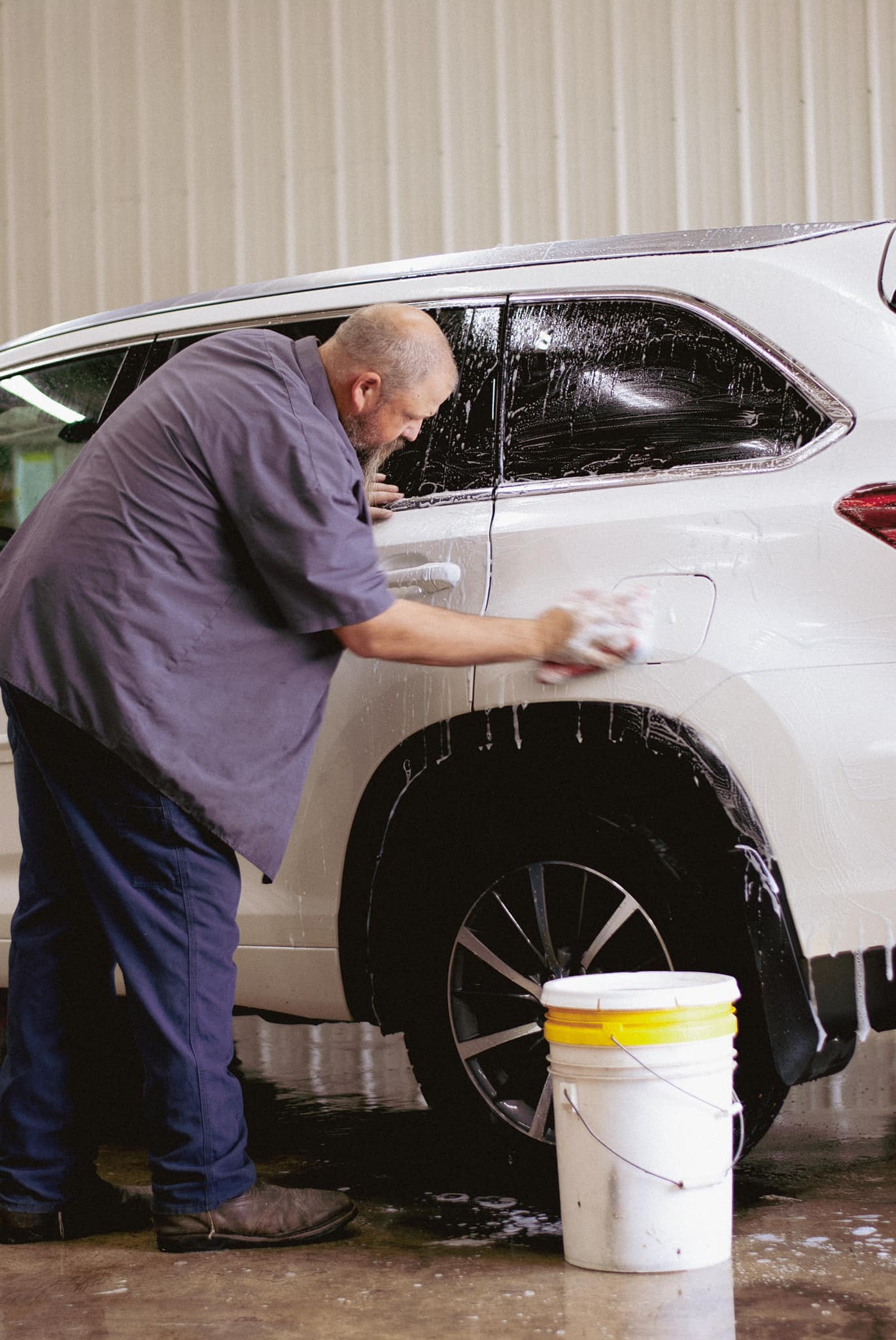 vehicle detailing services Man washing the back of a white SUV with a sponge while standing next to a yellow bucket in a garage.