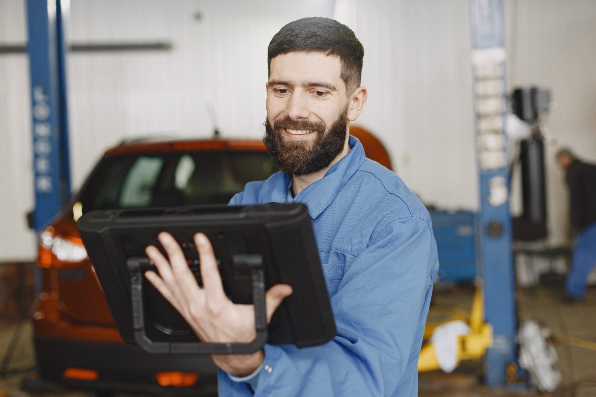 Get in Touch With Mid-Illini Collision Today A mechanic in blue coveralls uses a diagnostic tablet in an auto repair shop with a car in the background.
